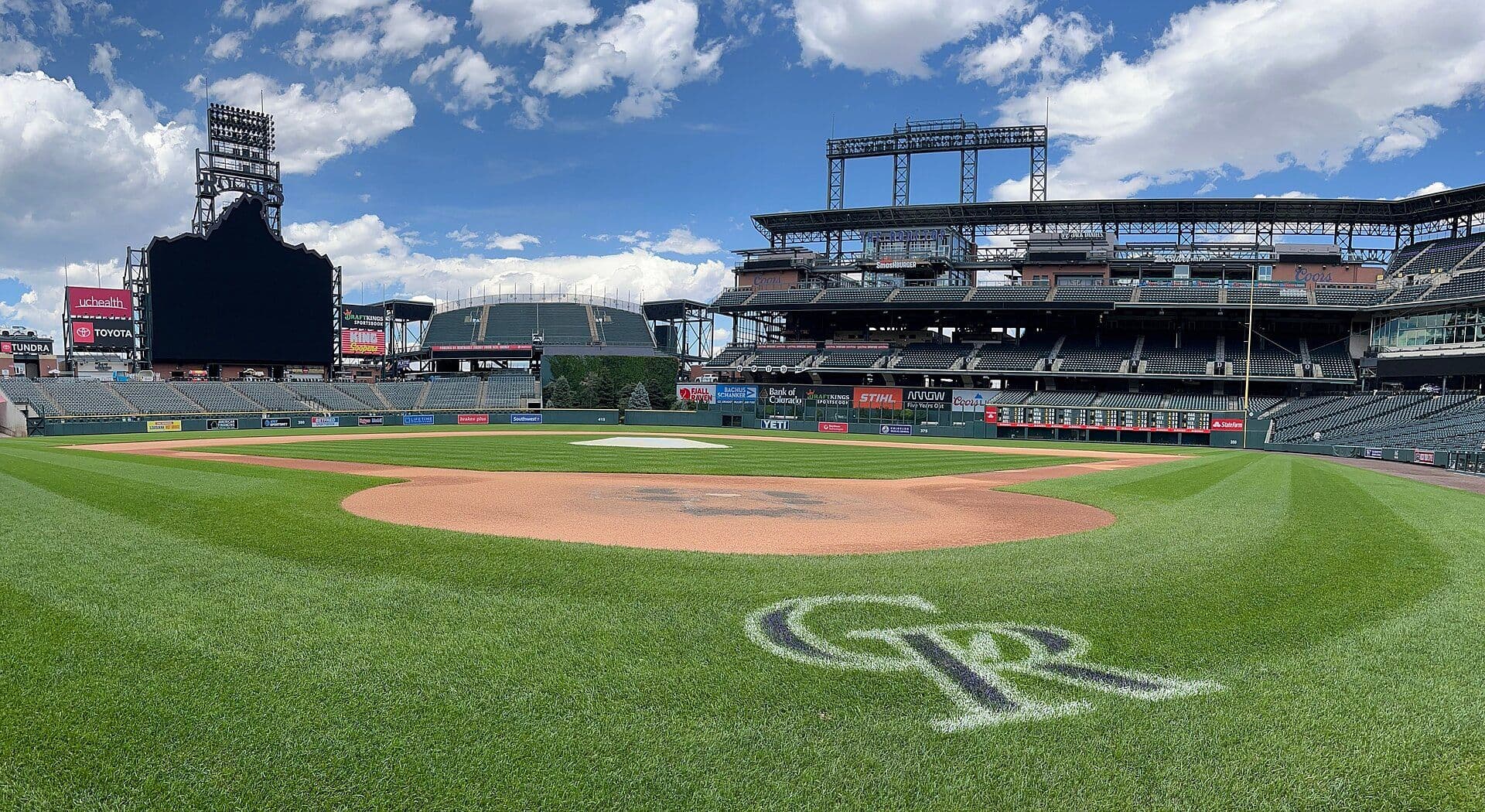 Field-level view of Coors Field in Denver from behind home plate, the Rockies logo on the grass under a blue sky