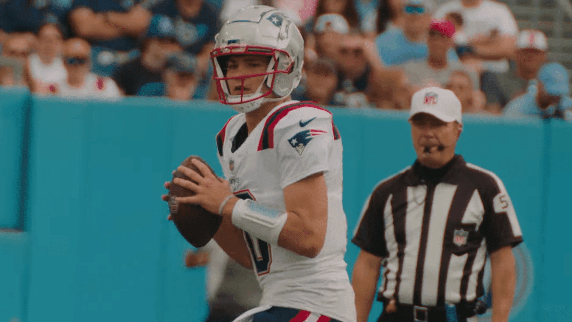 New England Patriots quarterback Drake Maye in a white road uniform holding the ball in the pocket, an official in the background.