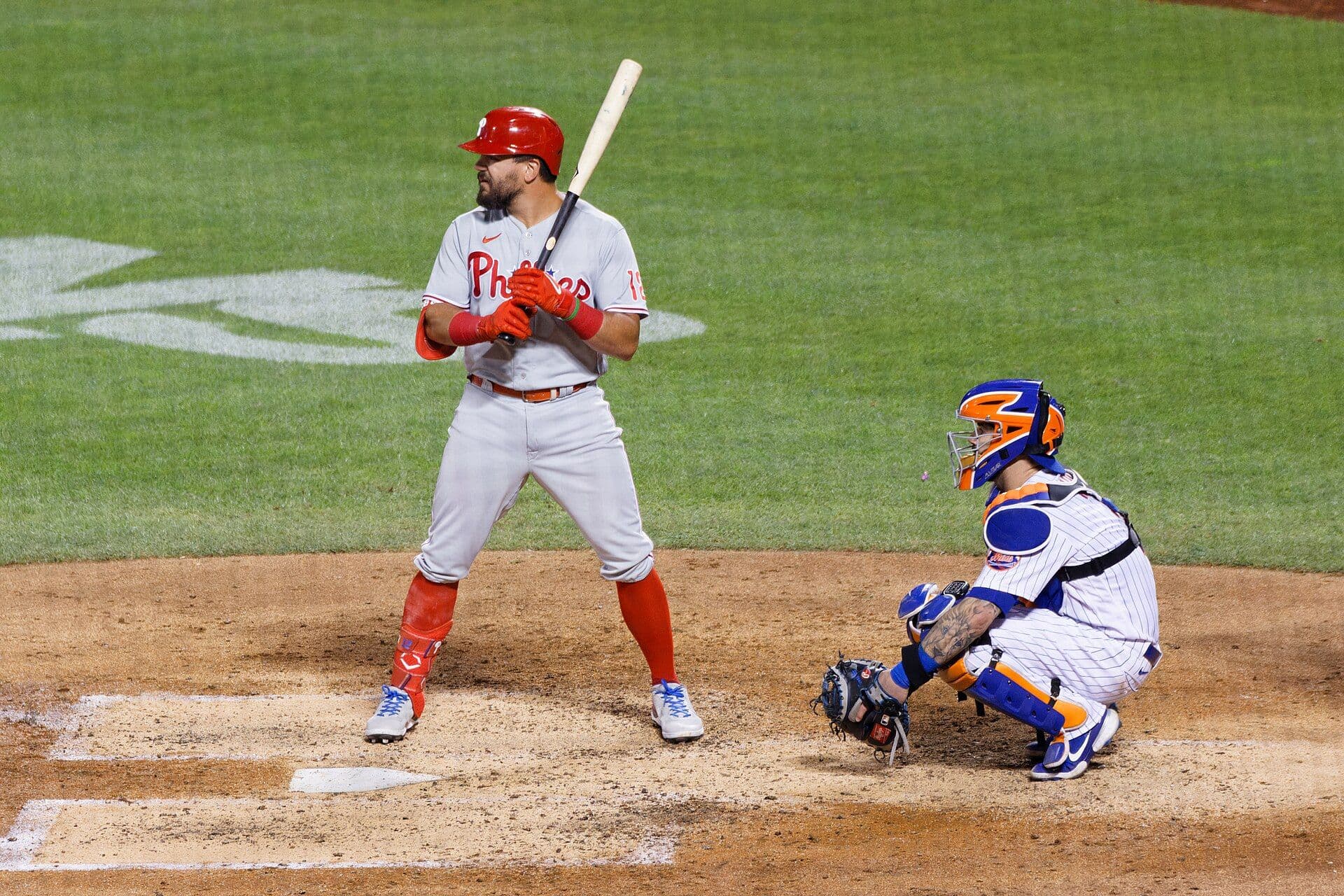 Kyle Schwarber of the Philadelphia Phillies stands in the batter's box in a gray Phillies road uniform and red helmet, bat held up, with a catcher crouched behind him.