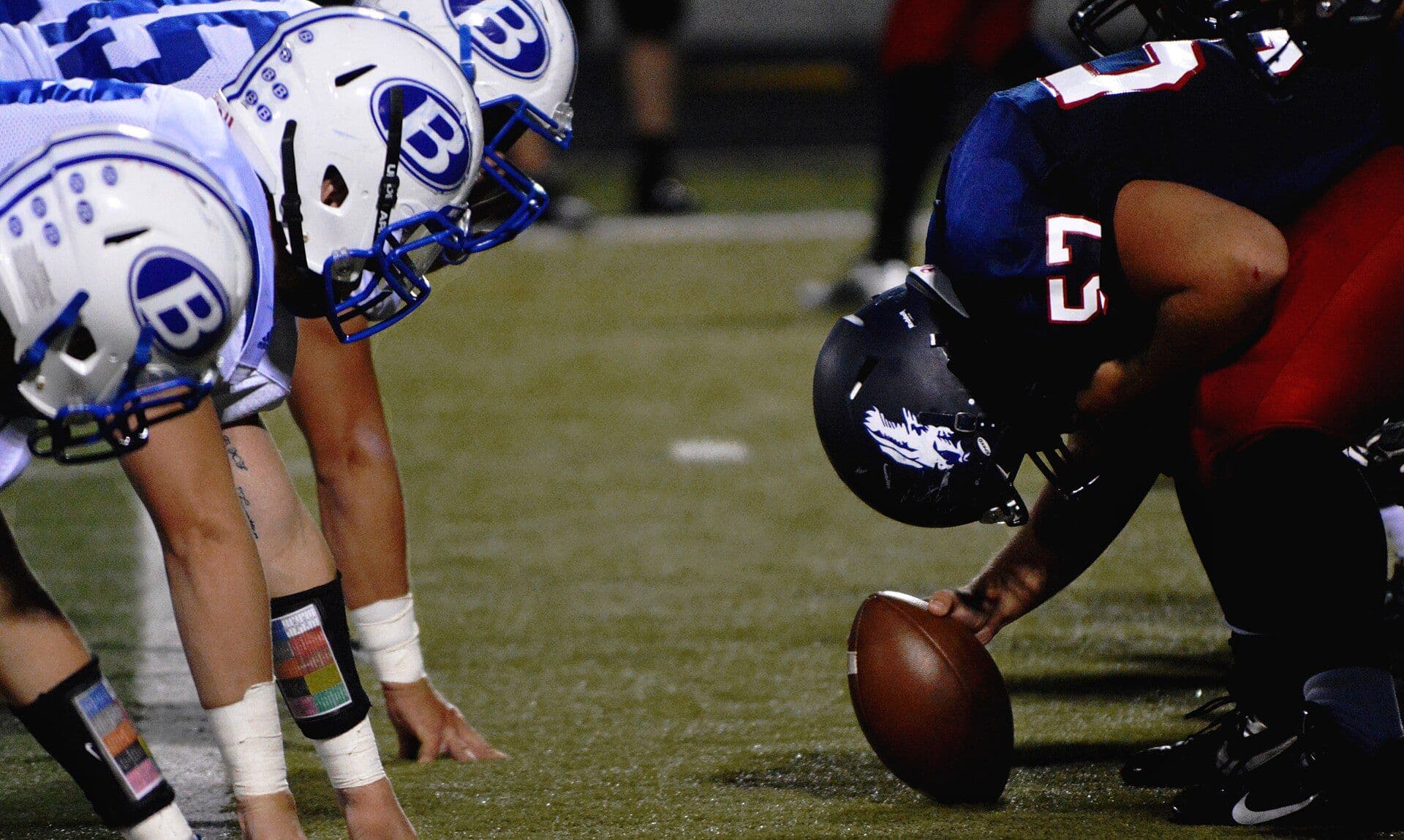 Two football lines set at the line of scrimmage under stadium lights at night, the center's hand on the ball, seen from field level.