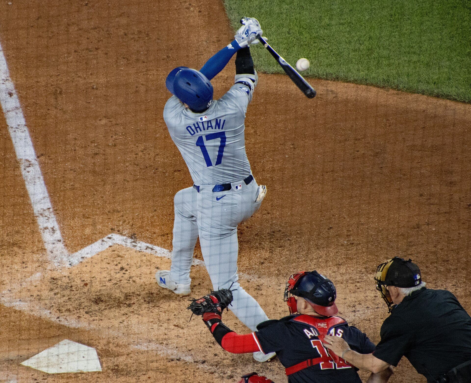 Shohei Ohtani follows through on a swing in a gray Los Angeles Dodgers road uniform, the name OHTANI and number 17 visible on his back, with the catcher and home plate umpire behind him.