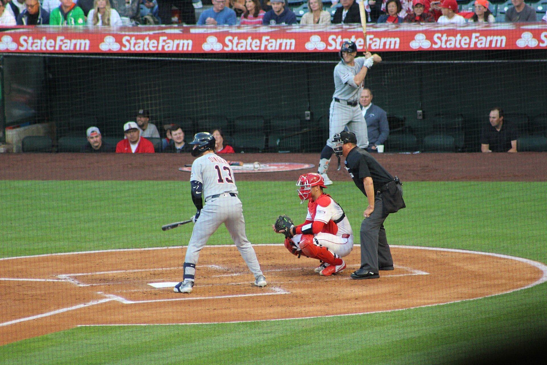 Dillon Dingler waits in the batter's box in a gray Detroit Tigers road uniform, the name DINGLER and number 13 visible on his back, with the catcher and home plate umpire set behind him.