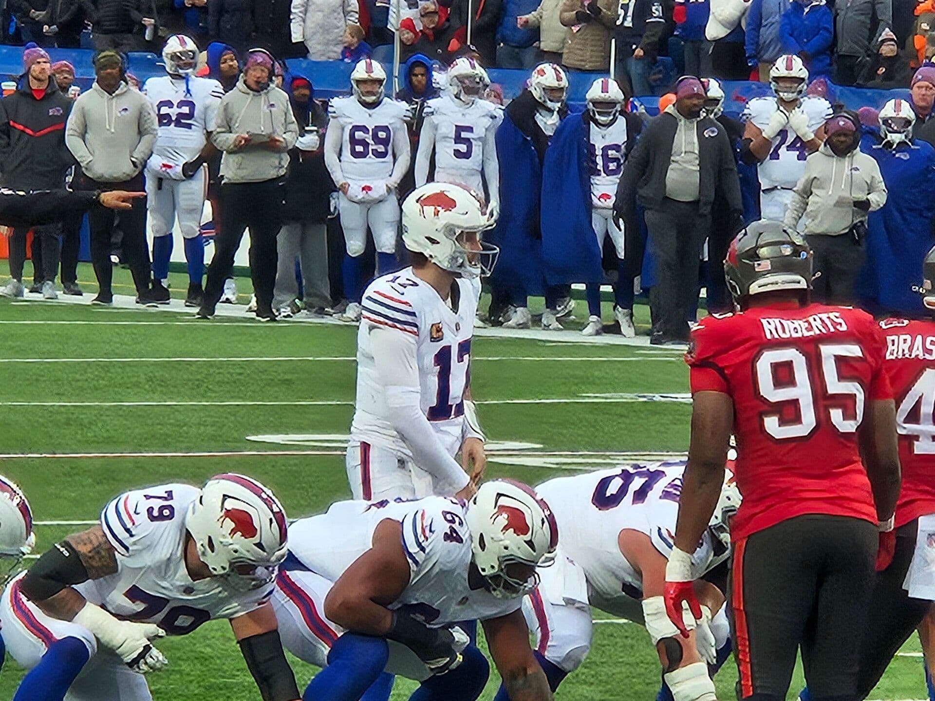 Buffalo Bills quarterback Josh Allen, number 17 in the white uniform, stands behind his offensive line before a snap with the Bills sideline in the background.